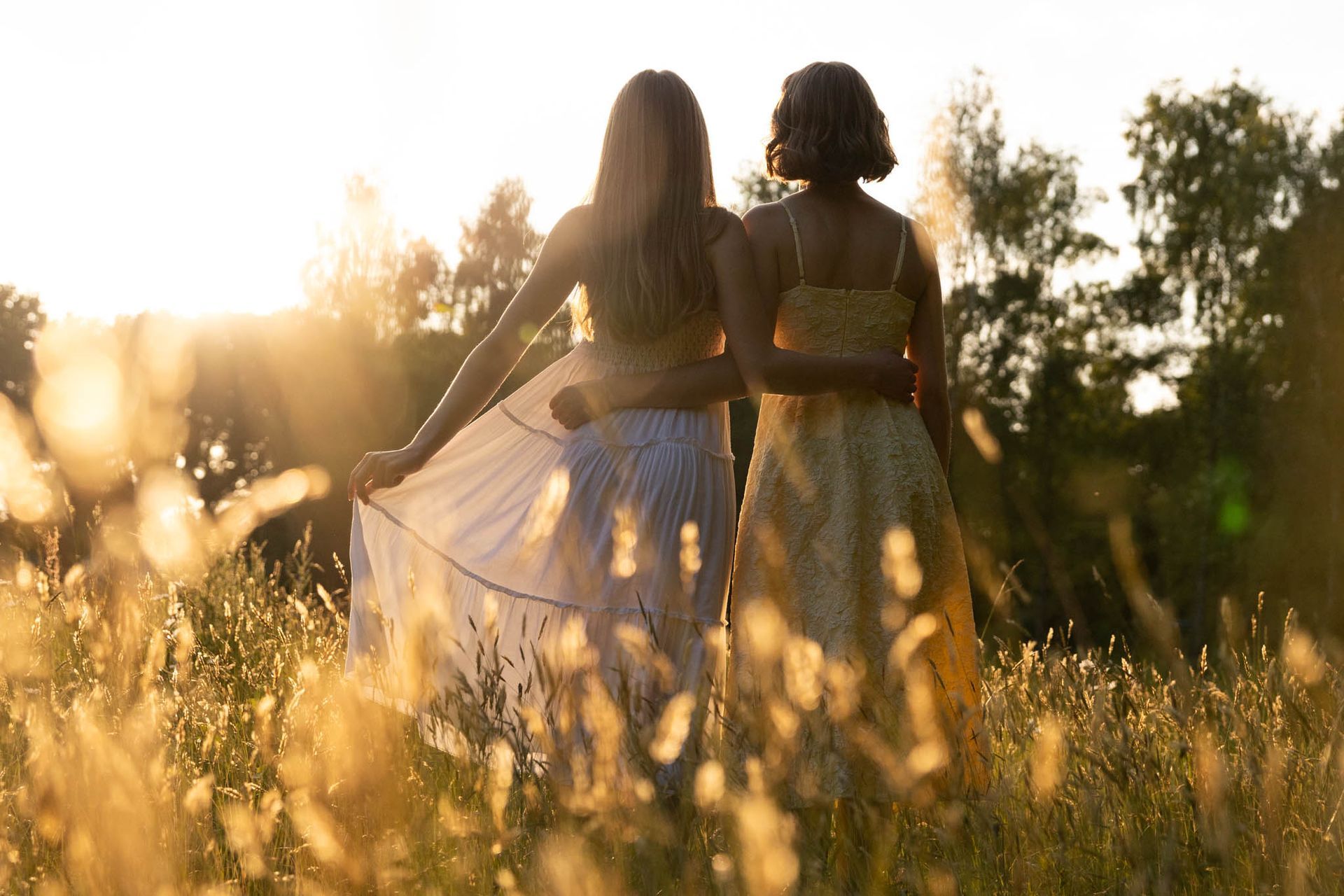 Twee mensen staan arm in arm in een veld met hoog gras, met hun rug naar de camera, terwijl de zon achter hen ondergaat.