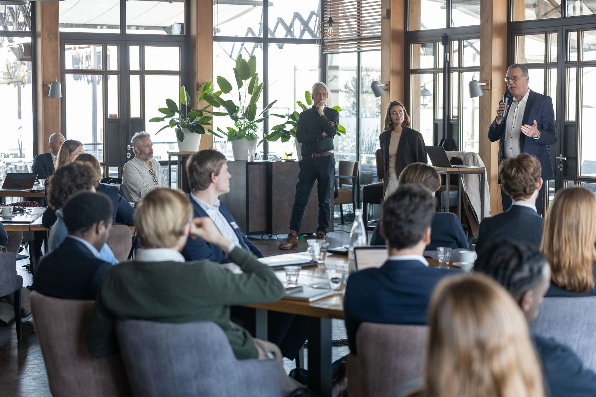 Mensen die een presentatie bijwonen in een kamer met houten lambrisering. Sprekers op het podium, publiek aan tafels.