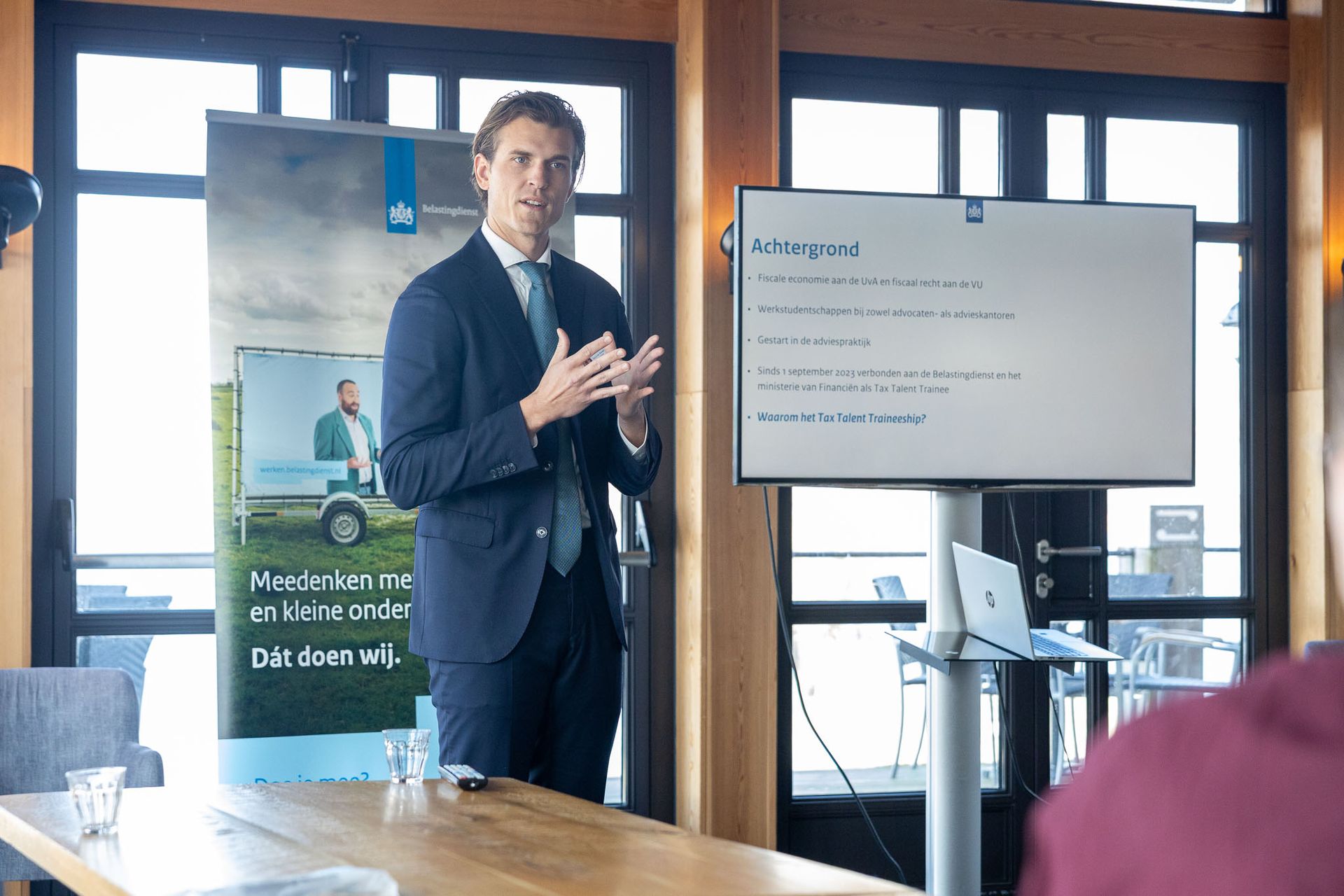Man in pak presenteert voor een whiteboard. Banner en laptop zichtbaar. Houten interieur.