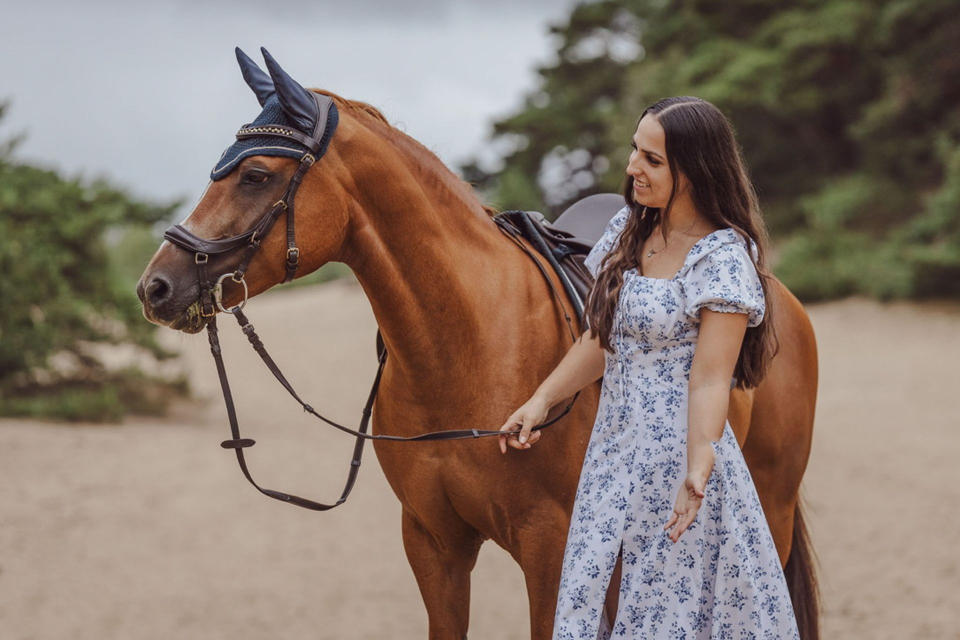 Een vrouw in een blauwe jurk lacht naar een bruin paard met rijuitrusting in de buitenlucht.