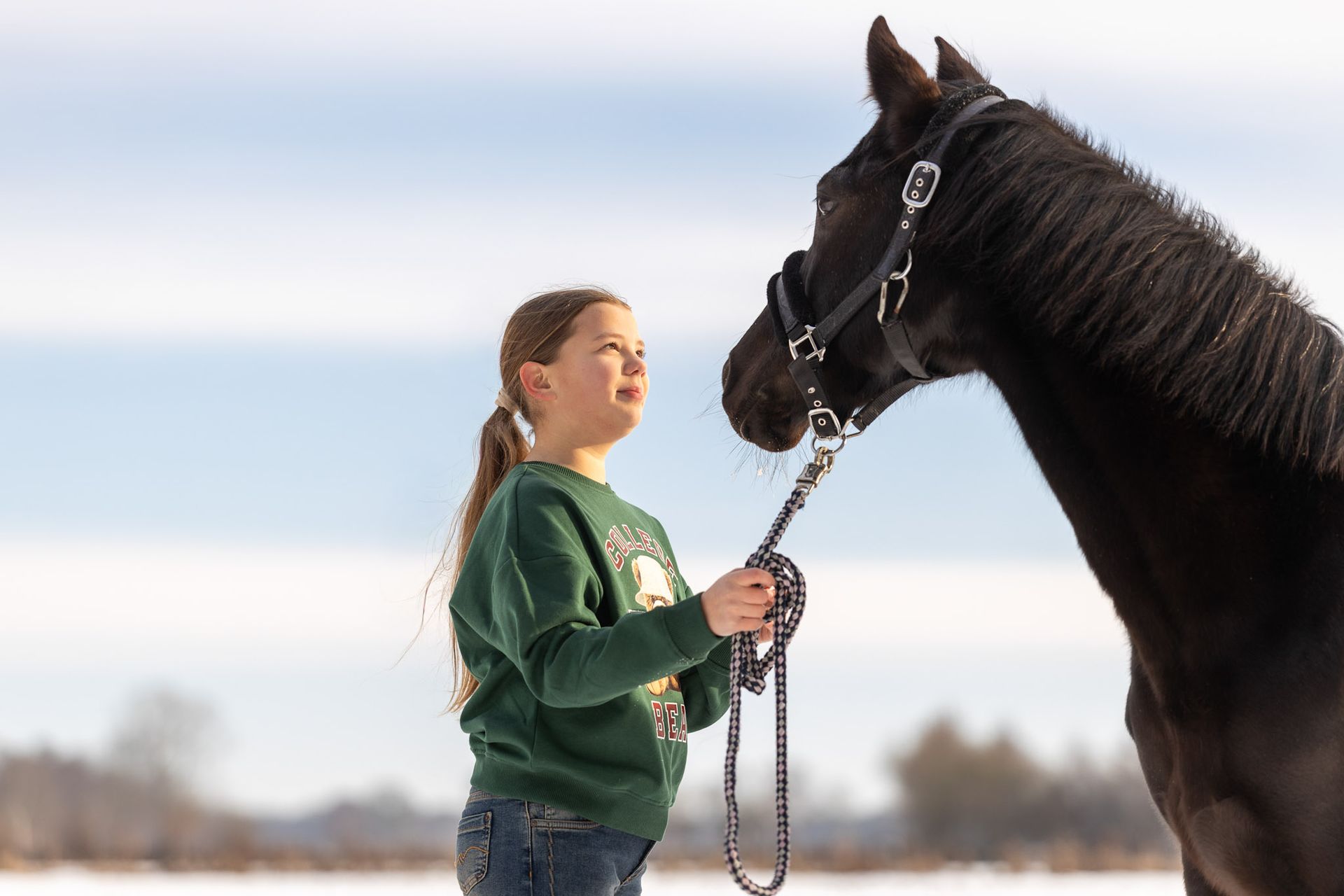 Een meisje in een groene trui kijkt op naar een zwart paard dat aan het halstertouw hangt, in een besneeuwd veld.