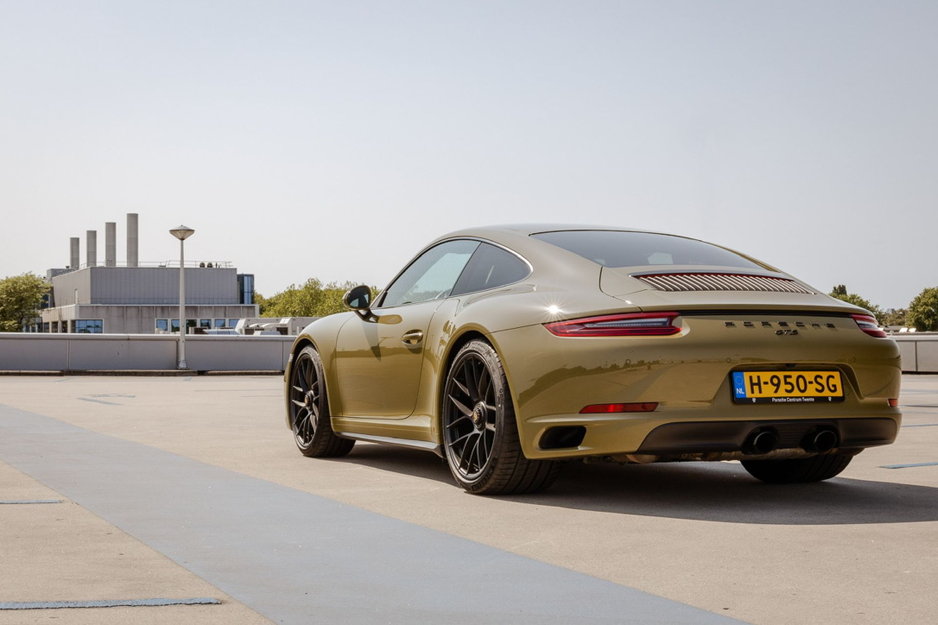 Een vrouw poseert naast een kaki Porsche sportwagen op een geplaveide weg.
