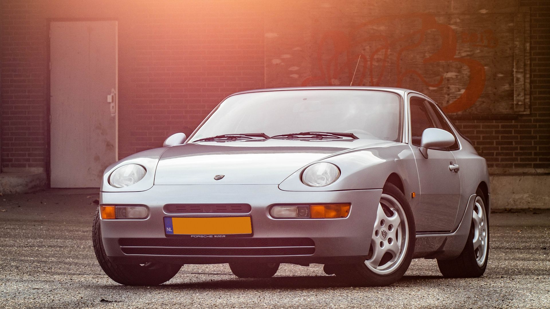Een zilverkleurige Porsche 968 coupé staat geparkeerd voor een bakstenen gebouw.
