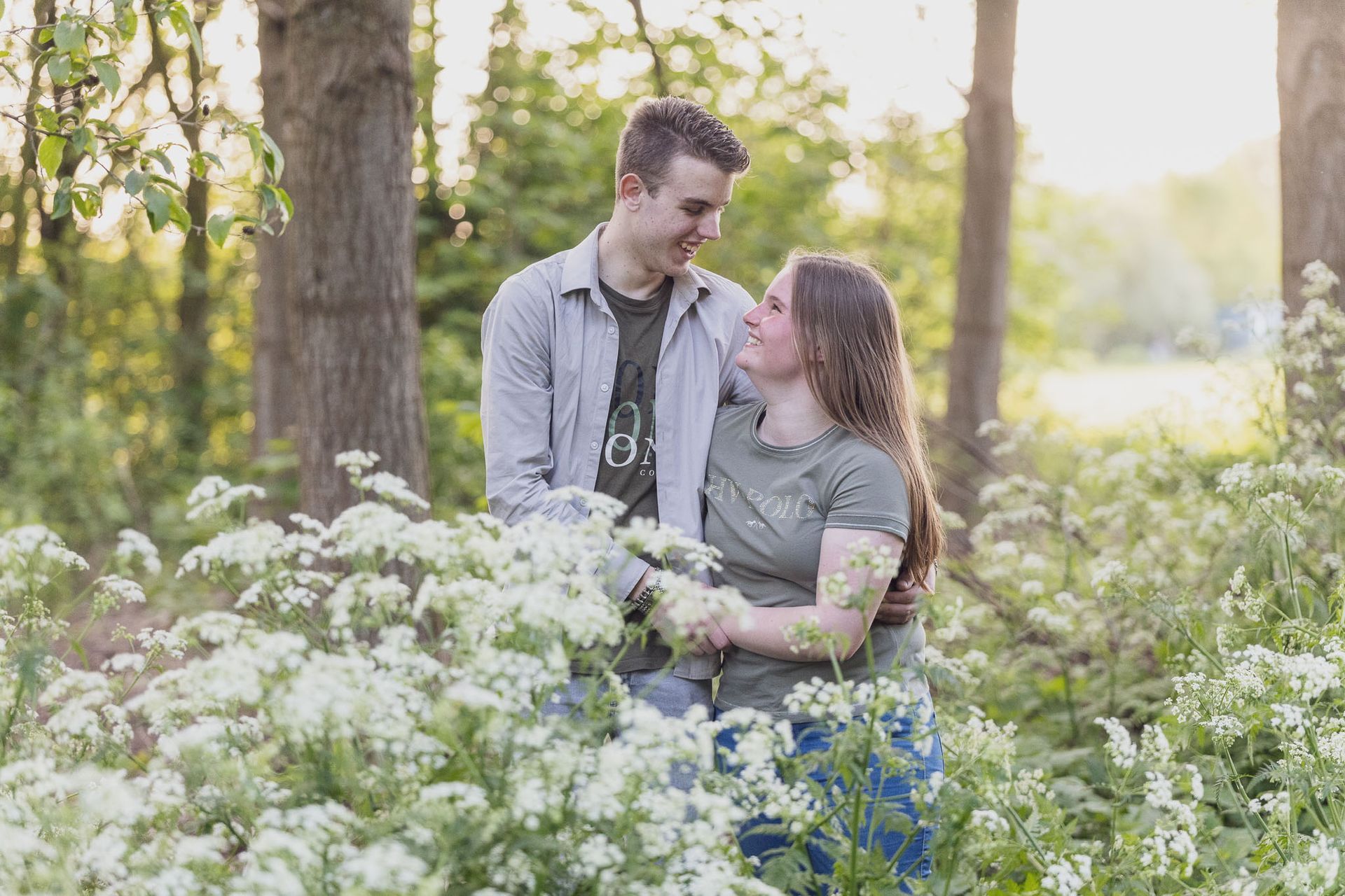 Een stel omhelst elkaar in een zonovergoten veld met witte bloemen en lacht elkaar toe.