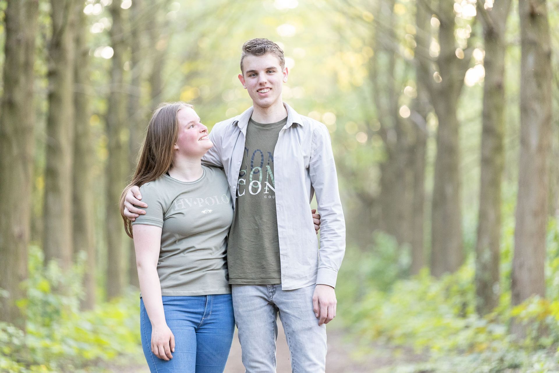 Een stel in een bos, de vrouw met haar arm om de taille van de man, glimlachend. Bomen op de achtergrond.
