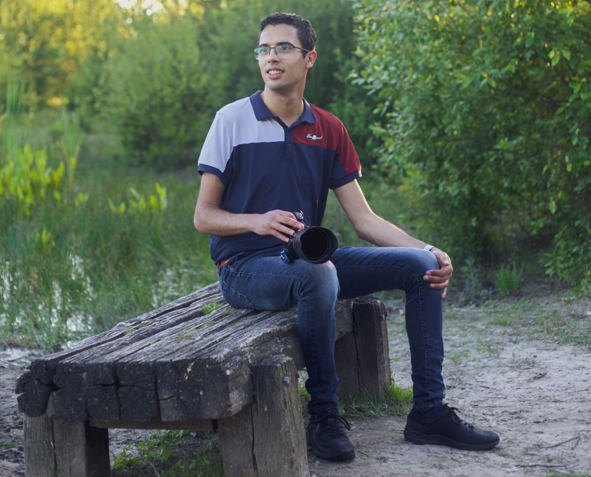 Een man in poloshirt en spijkerbroek zit op een houten bankje, houdt een cameralens vast en kijkt opzij, naar buiten.