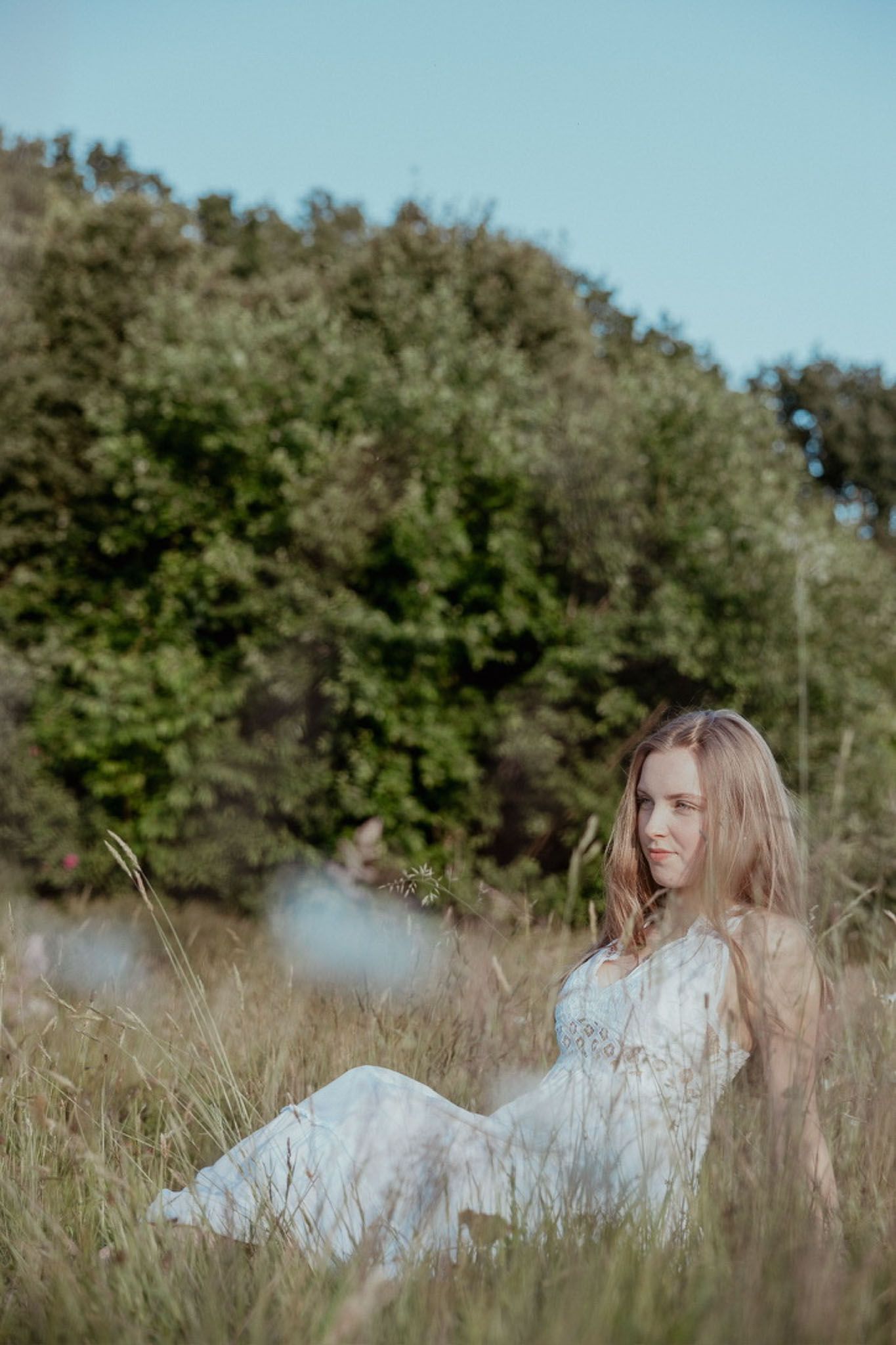 Vrouw in witte jurk ligt lachend in het hoge gras. Bomen en blauwe lucht op de achtergrond.