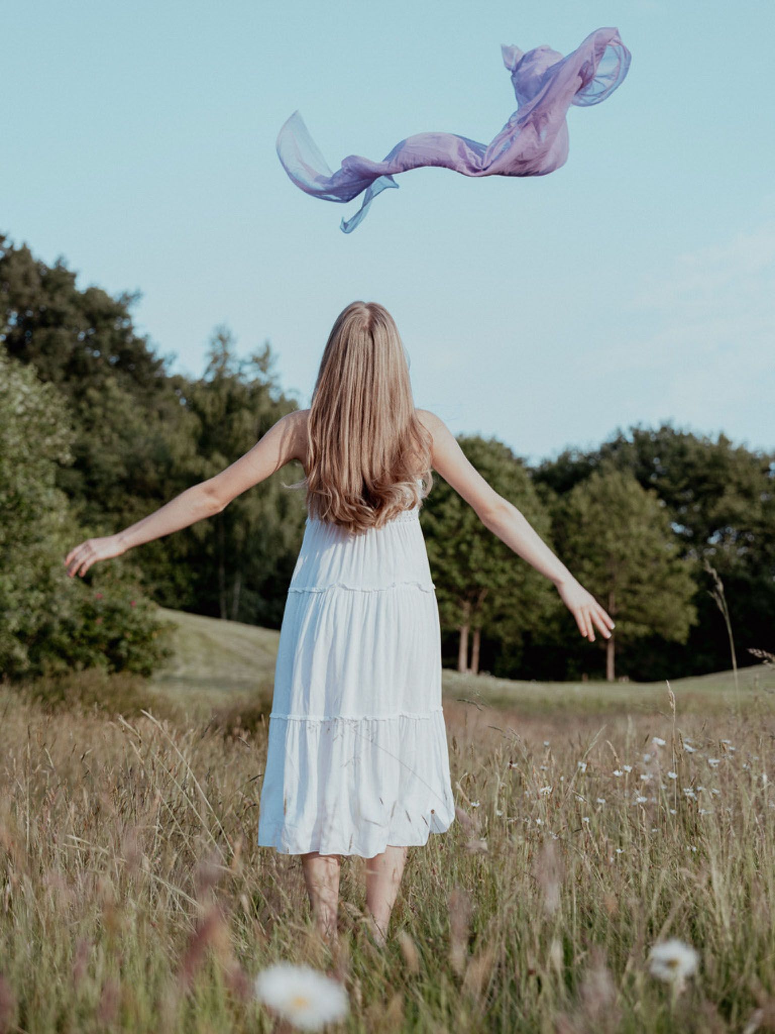 Vrouw in een witte jurk met uitgestrekte armen in een veld, sjaal wapperend boven haar, blauwe lucht.