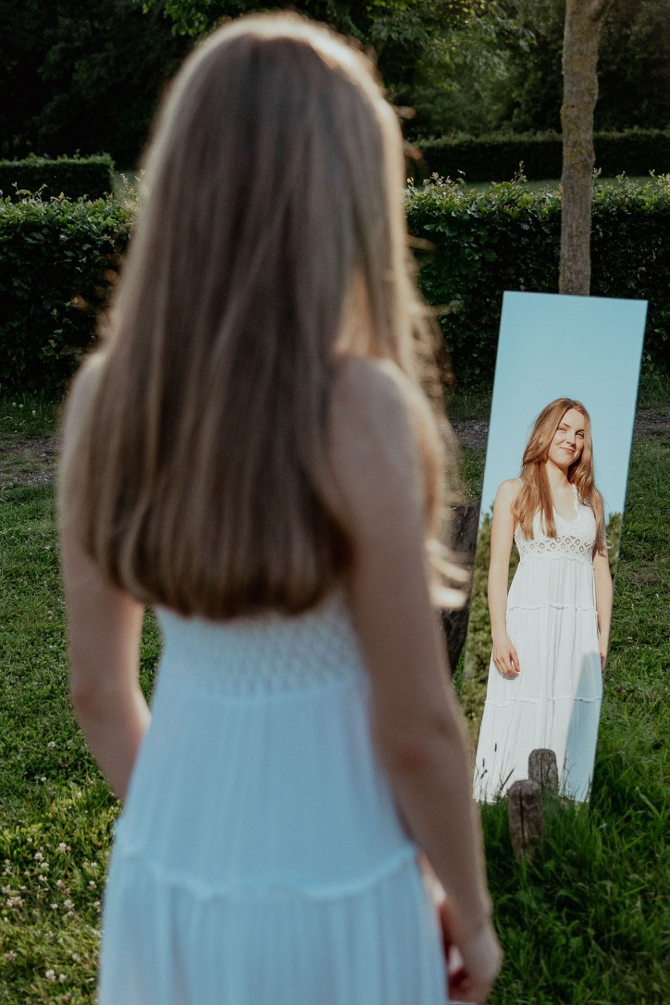 Vrouw in witte jurk kijkt buiten in de spiegel, op een zonnige dag.