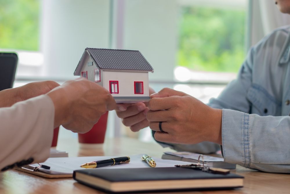 Hands passing a model house; paperwork, pen, and notebook on a table.