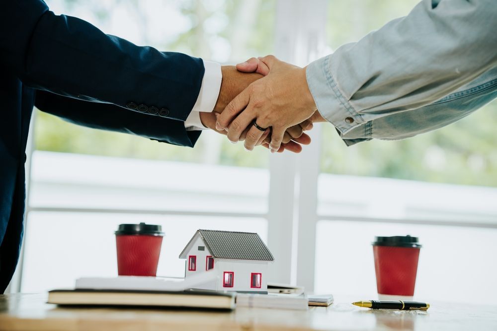 Two people shake hands over a table with a house model, coffee cups, and paperwork.