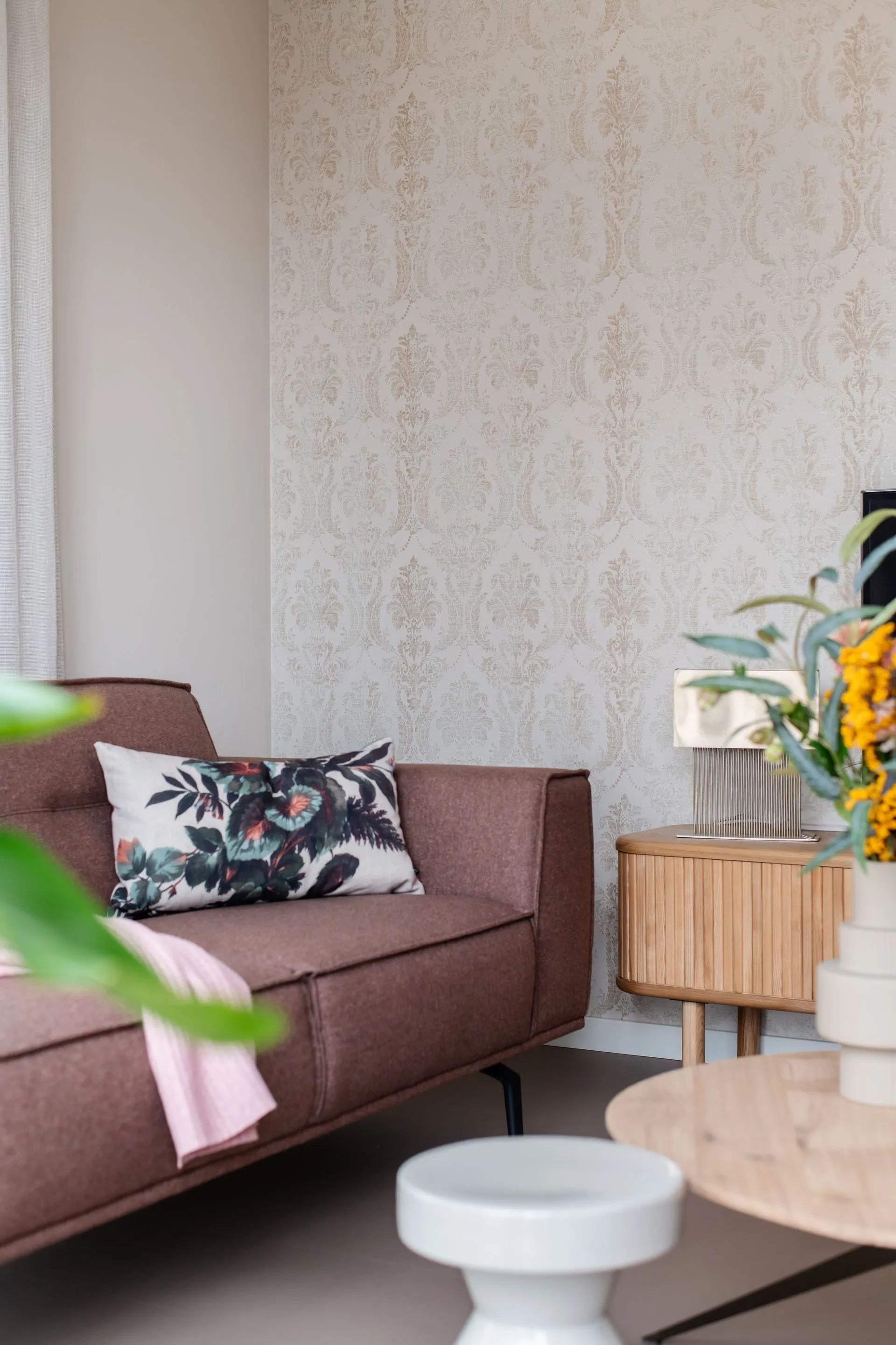 Woonkamer met een mauve bank, bloemenkussen, behang met patroon en een houten bijzettafel.