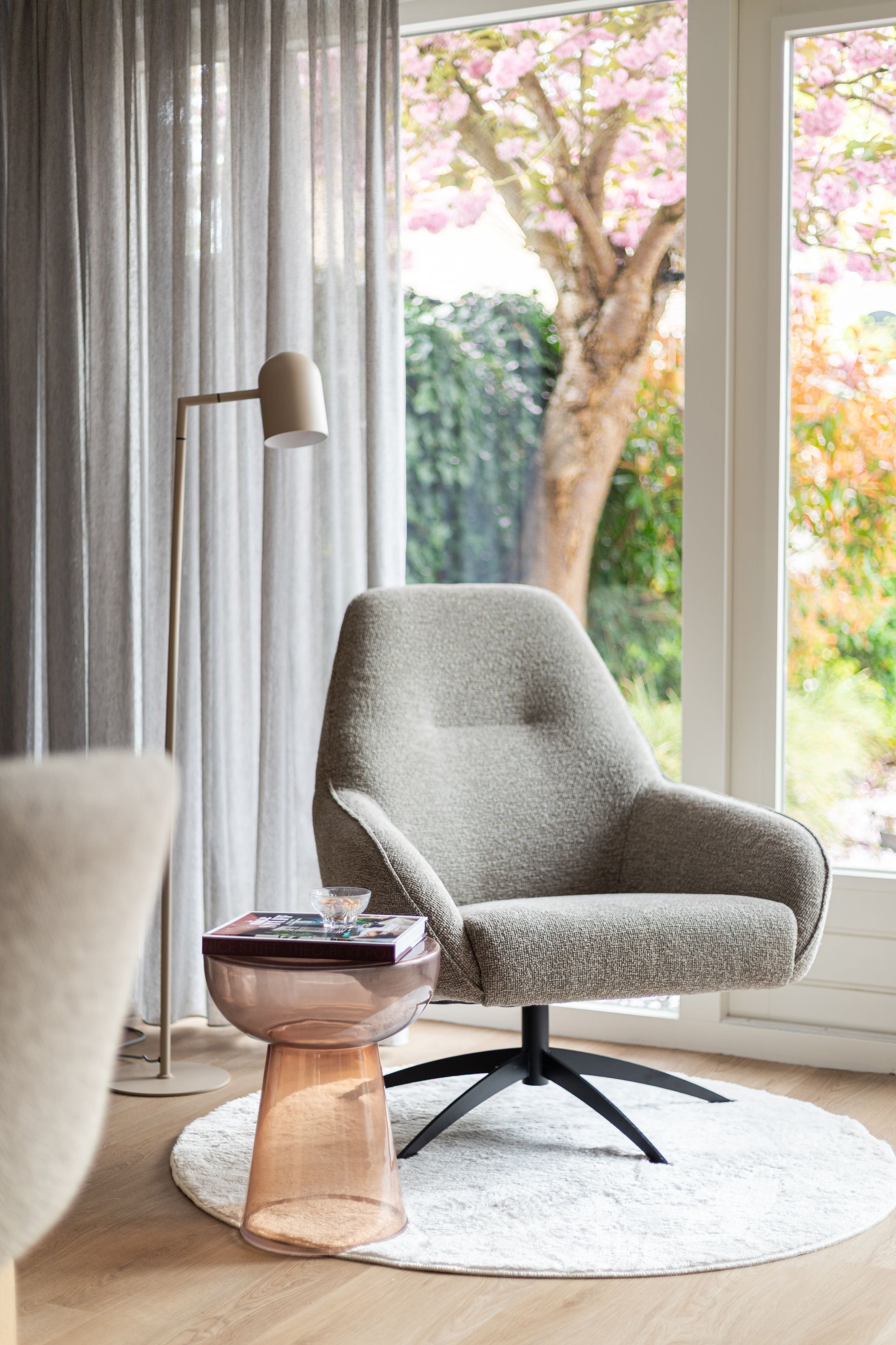 Gezellig leeshoekje: grijze draaistoel, gouden staande lamp, bijzettafel, raam met uitzicht op de boom.