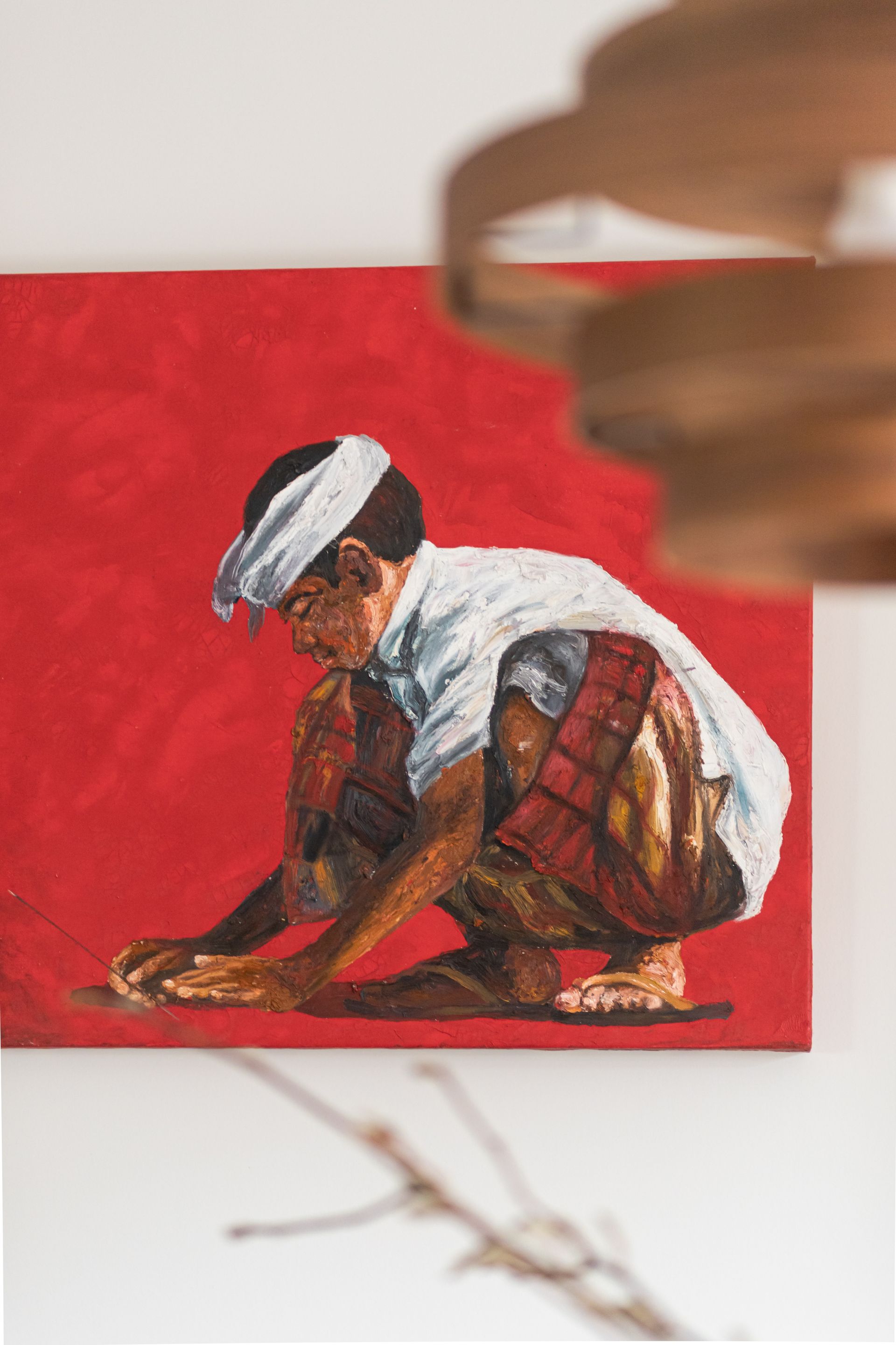 Schilderij van een gehurkte persoon voor een rode achtergrond. Hij draagt een wit shirt en een witte hoofdbedekking.