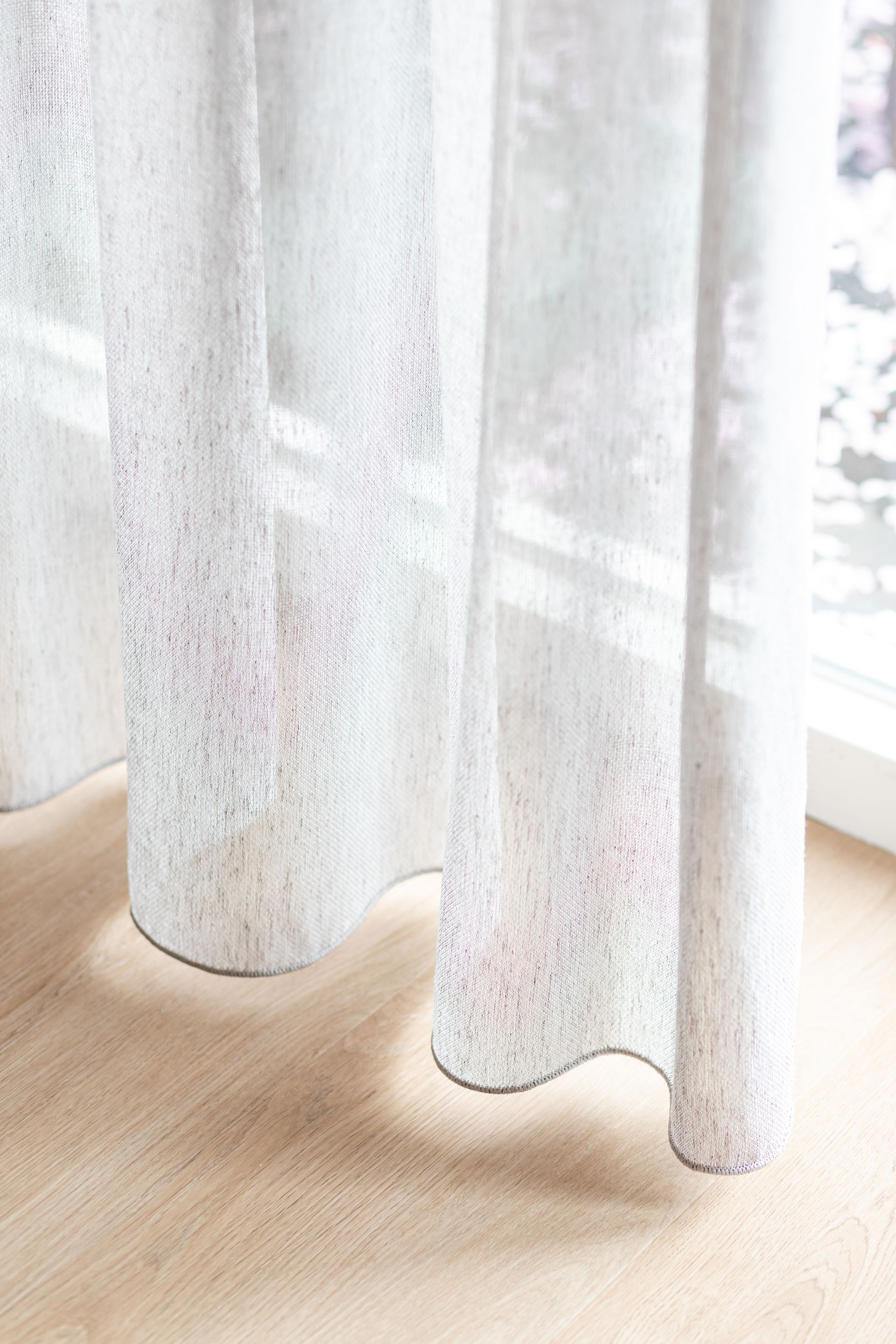 Witte, transparante gordijnen met een structuurpatroon hangen bij een raam en werpen schaduwen op de houten vloer.