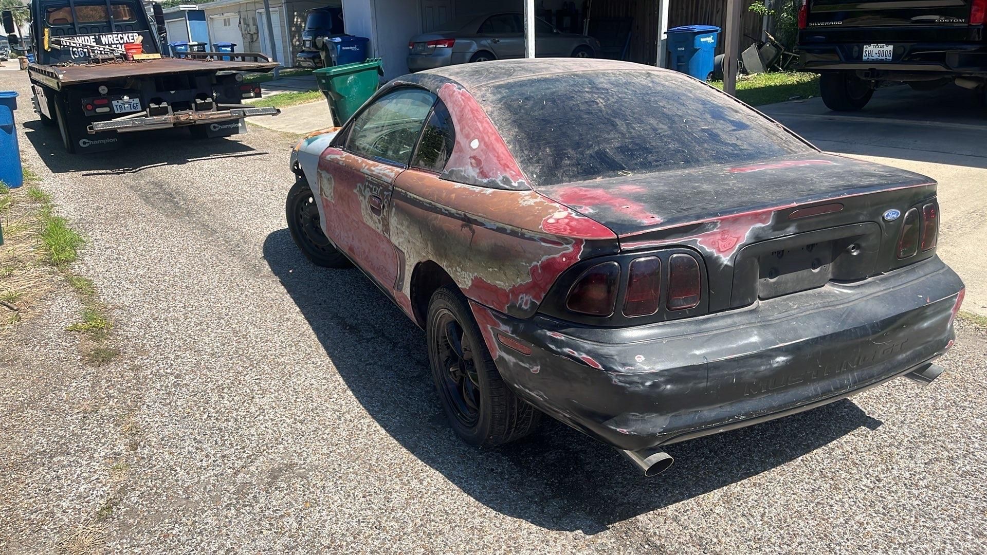 White car with hood open, front end missing, in a junkyard.
