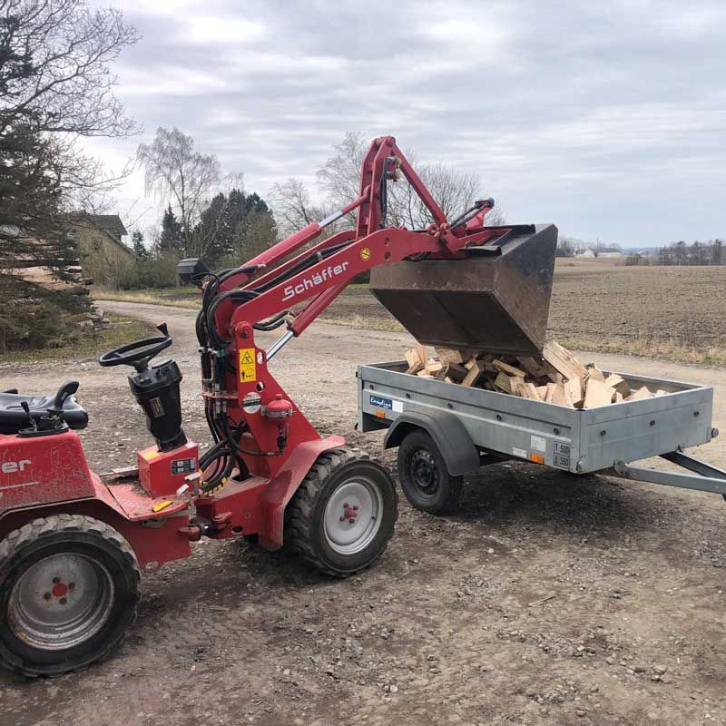 Rød Scheffer-læsser tømmer brænde ned i en lille trailer på en grusvej.