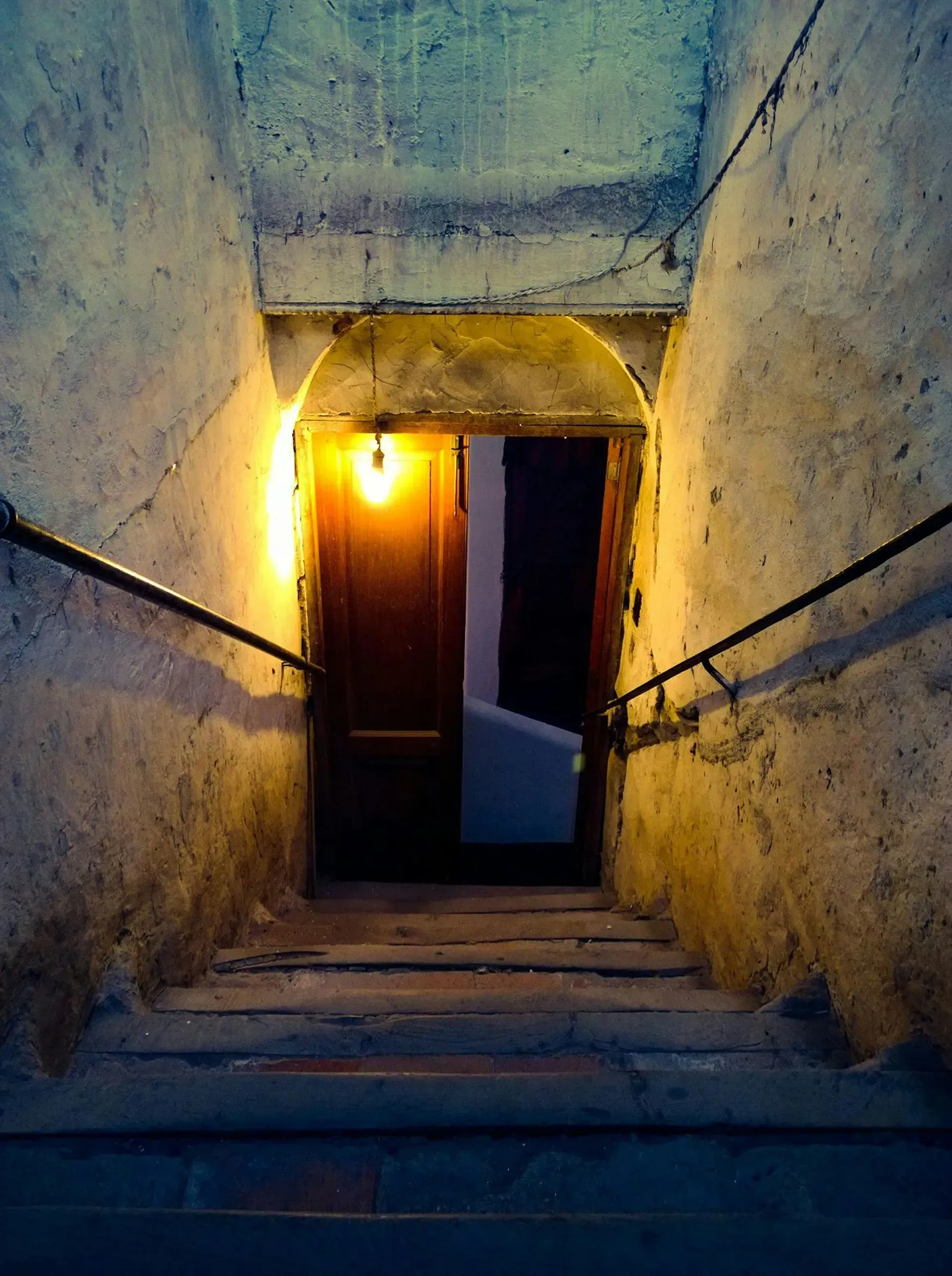 Narrow stone stairwell leading to a dim doorway, lit by a warm yellow lamp at the top