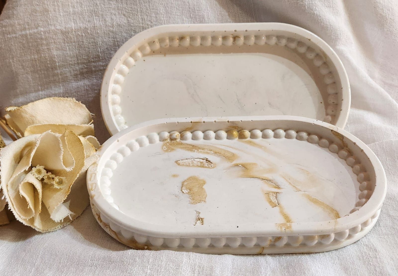 Dos jaboneras de cerámica blanca, de forma ovalada, una de ellas con manchas, y flores secas sobre un paño blanco.