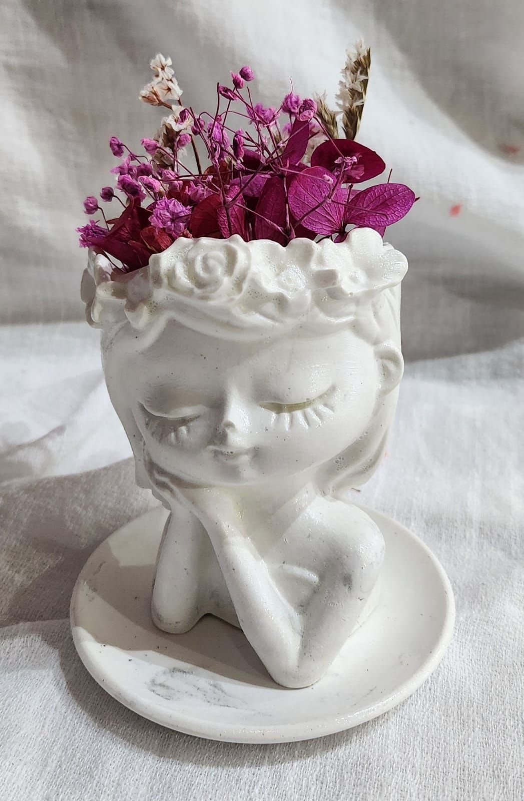 Maceta de cerámica blanca con forma de cabeza de mujer, que contiene flores en un pequeño plato.