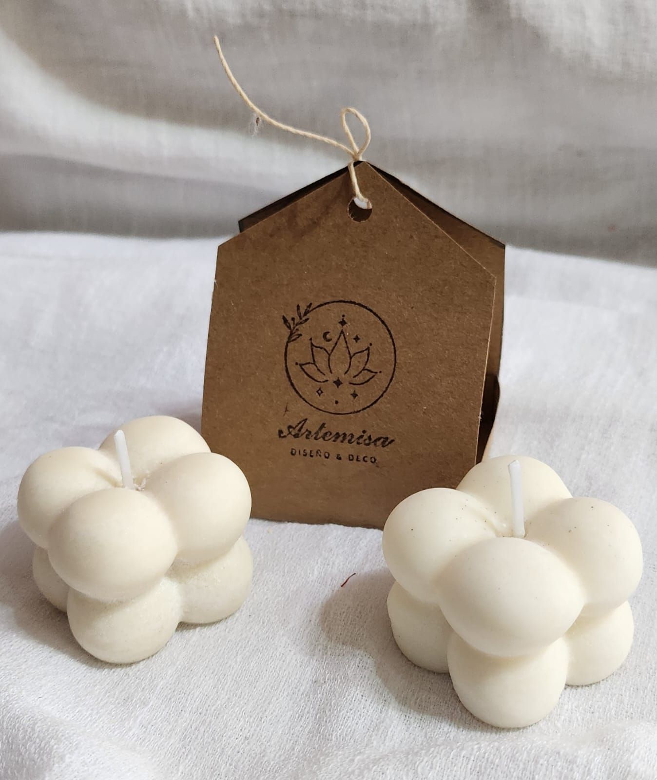 Dos velas blancas de burbujas y una etiqueta marrón con el logotipo sobre un paño blanco.