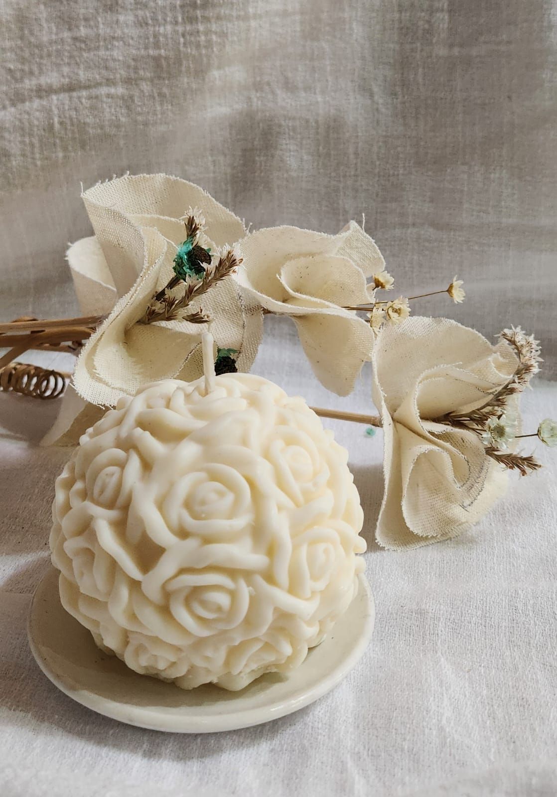 Vela blanca en forma de rosa sobre un pequeño plato, con decoraciones florales delante de un fondo texturizado.