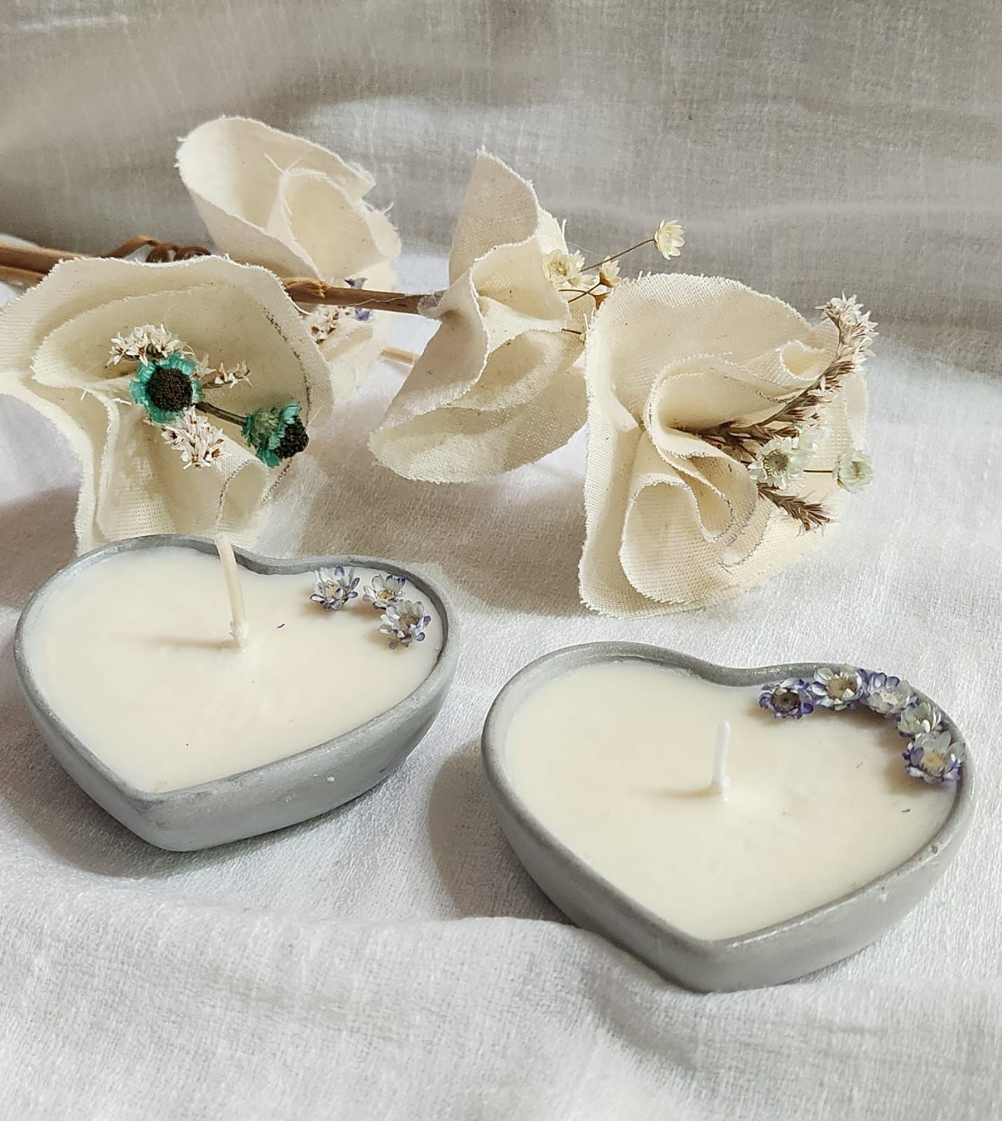 Dos velas en forma de corazón en recipientes grises, decoradas con flores secas, con flores de color claro al fondo.