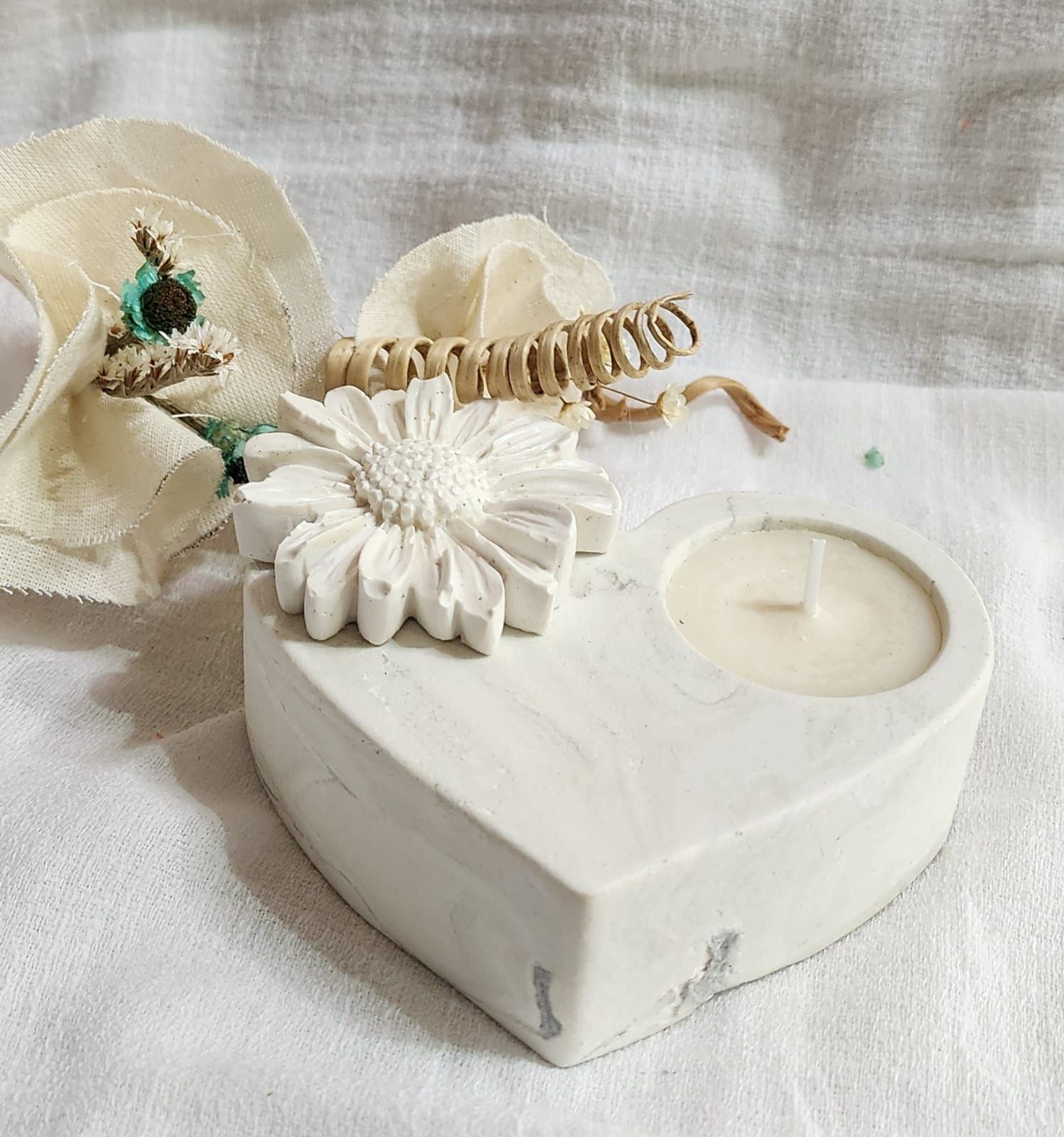 Portavelas de hormigón blanco en forma de corazón con una flor y una vela encendida sobre una superficie de tela blanca.