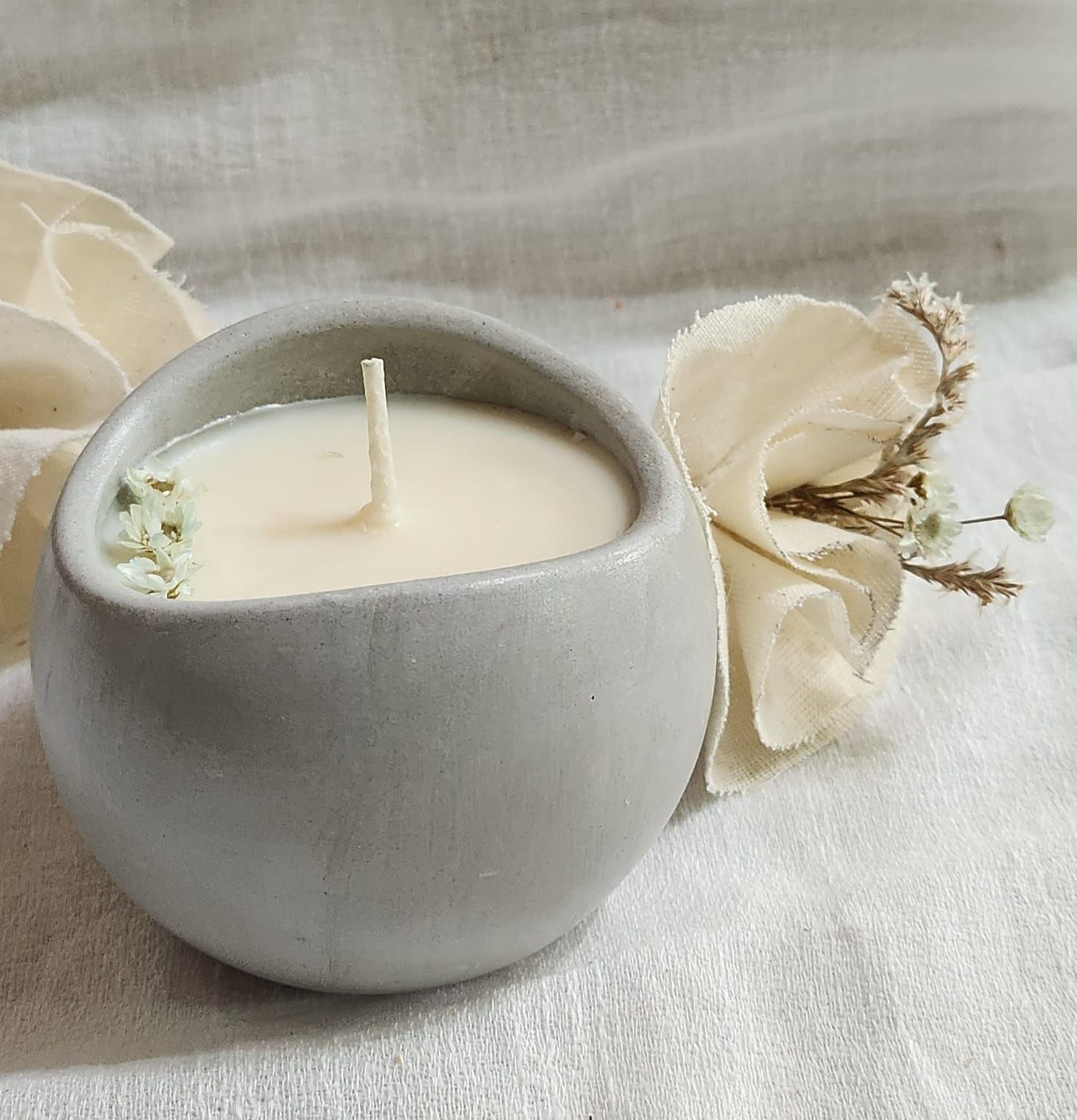 Una vela redonda gris en un recipiente de hormigón con una mecha blanca, decorada con flores secas sobre un paño blanco.