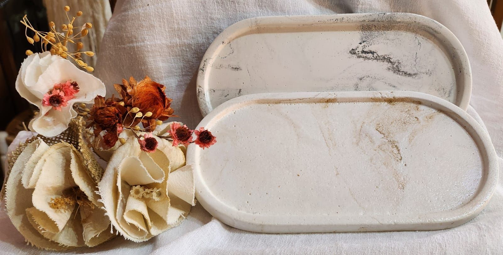 Dos bandejas ovaladas de hormigón, una con vetas grises, adornadas con flores sobre una toalla blanca.