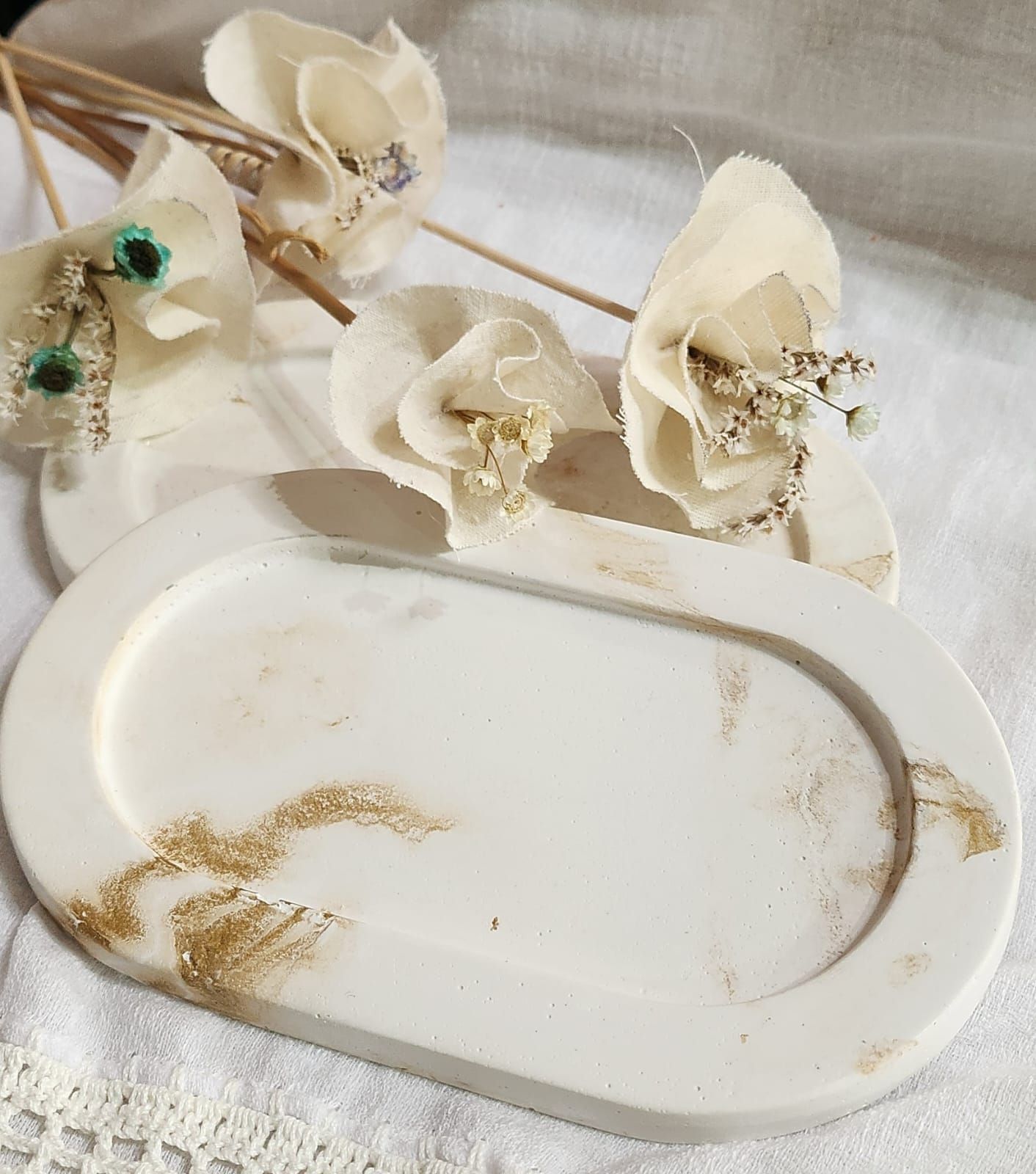 Dos bandejas ovaladas blancas con detalles dorados, coronadas con decoraciones florales color crema, sobre un mantel blanco.