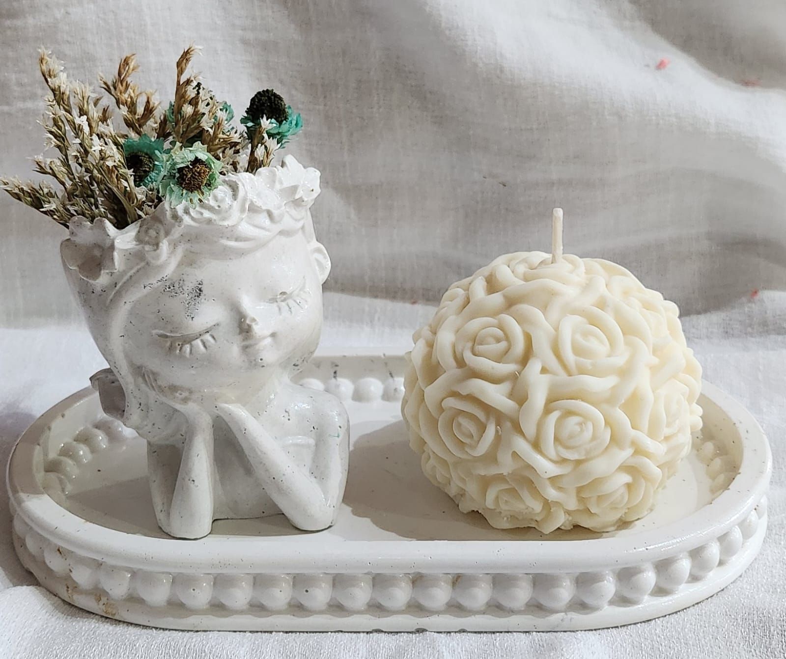 Maceta blanca con forma de ángel, flores secas y una vela con forma de rosa sobre una bandeja ovalada.