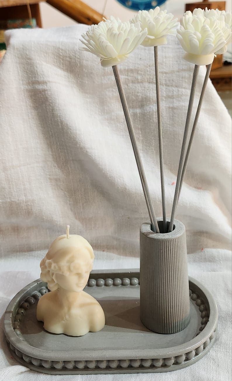 Bandeja gris con una vela en forma de busto y un difusor de caña con flores blancas.
