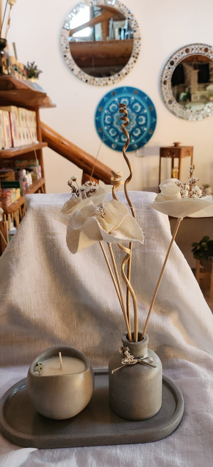 Una vela gris y un jarrón sobre una bandeja con flores, sobre una mesa con adornos en la pared al fondo.