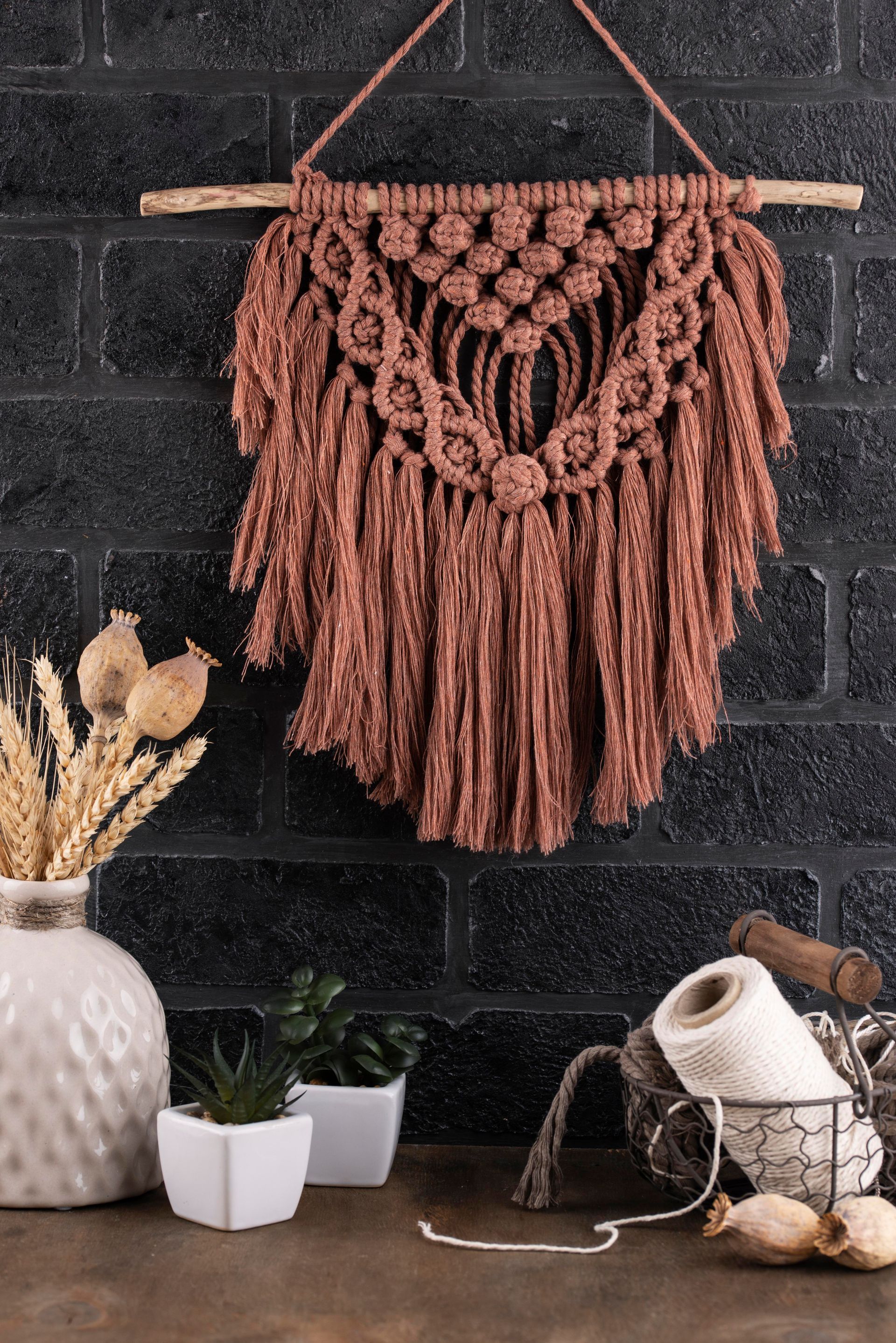 Tapiz de macramé marrón sobre una pared de ladrillo oscuro con elementos decorativos sobre una superficie de madera.