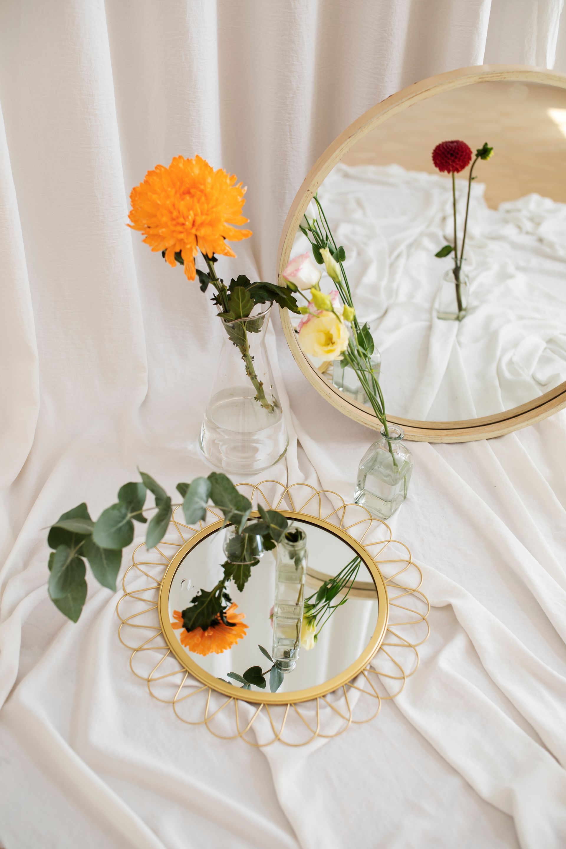 Flores naranjas y rojas en jarrones sobre un mantel blanco, reflejadas en dos espejos redondos.