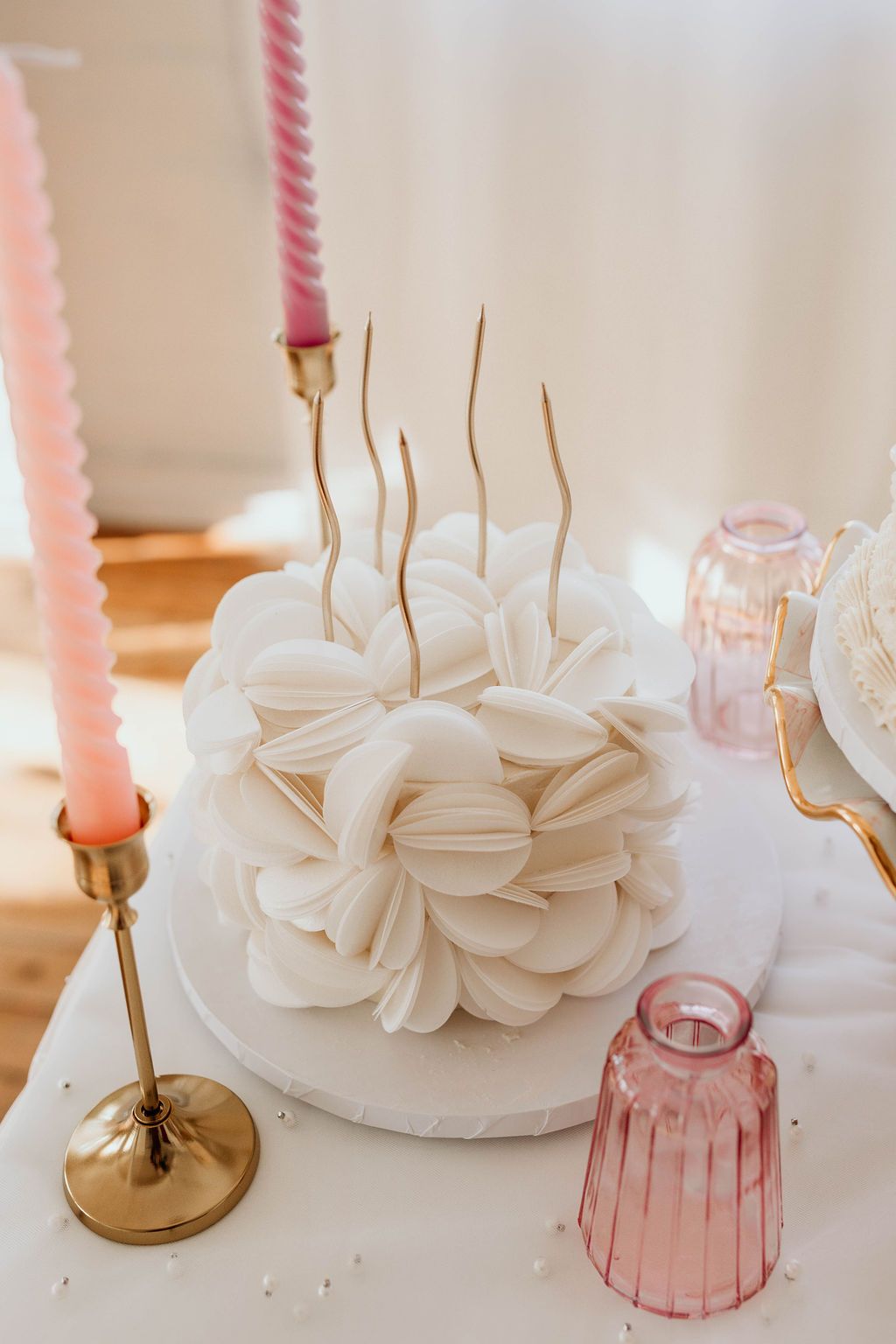 A textured white cake adorned with gold wire accents sits on a white platter, flanked by two pink candles and a vase.