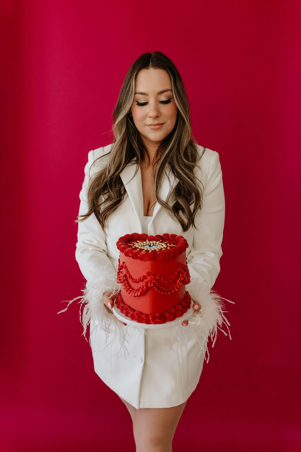 A person in a white, feather-trimmed blazer holds a red heart-shaped cake against a solid red background.