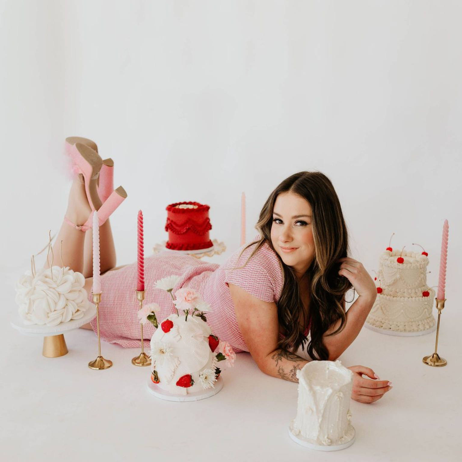 A person in a pink dress lies on a white floor surrounded by several small decorated cakes and pink tapered candles.