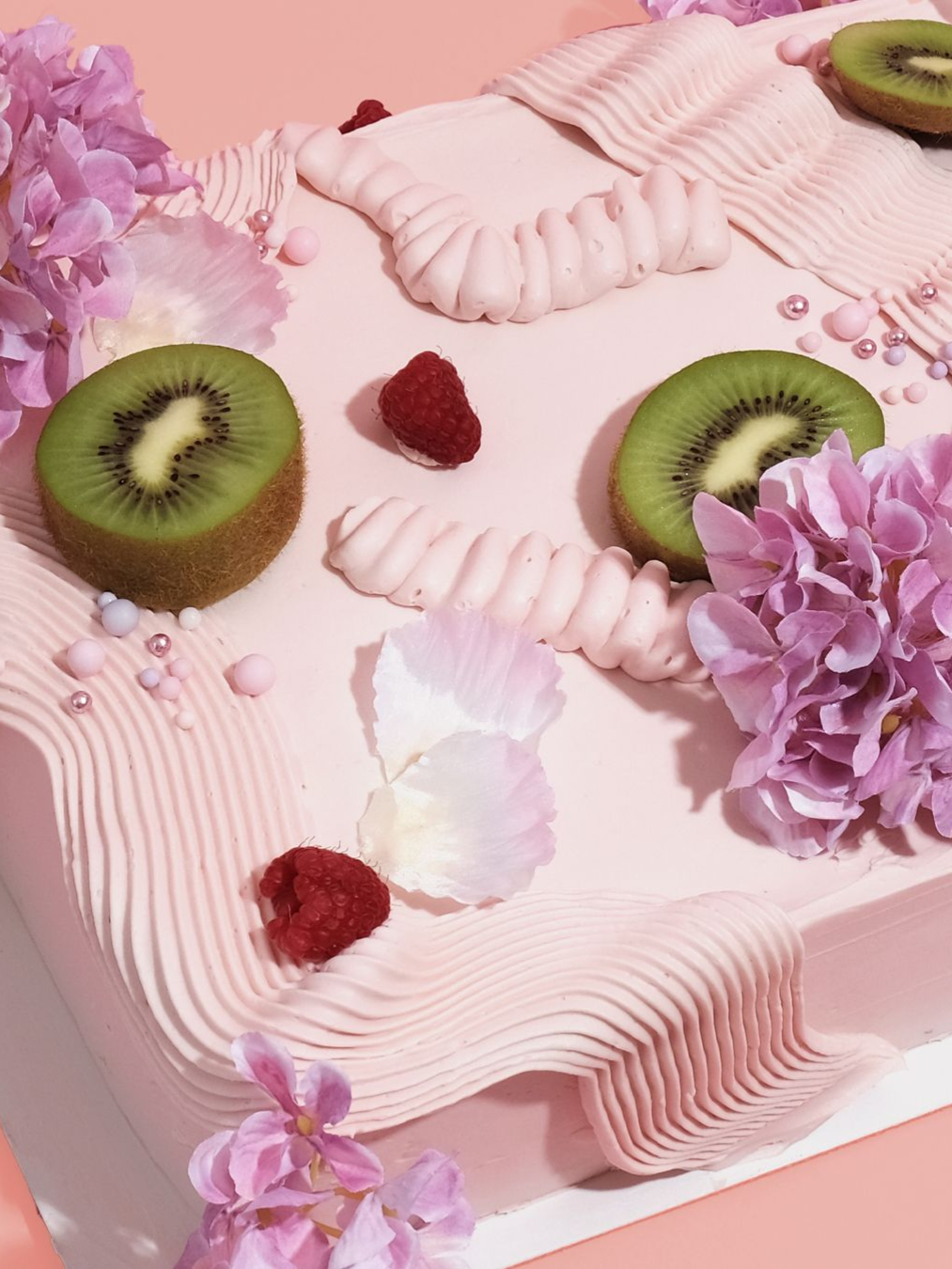 A square pink cake decorated with fresh kiwi slices, raspberries, purple flowers, and piped frosting ribbons.