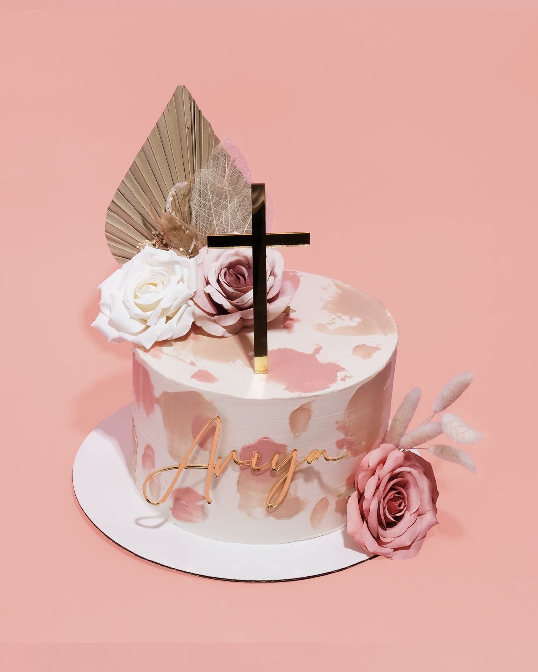 A cake decorated with pink splotches, roses, dried leaves, and a cross topper, with the name 