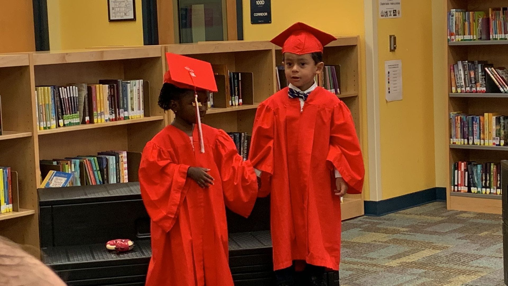 Two children in red graduation gowns are standing in a library