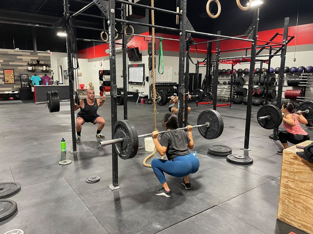 People squatting with barbells in a gym. Black floor, weights, and workout equipment.