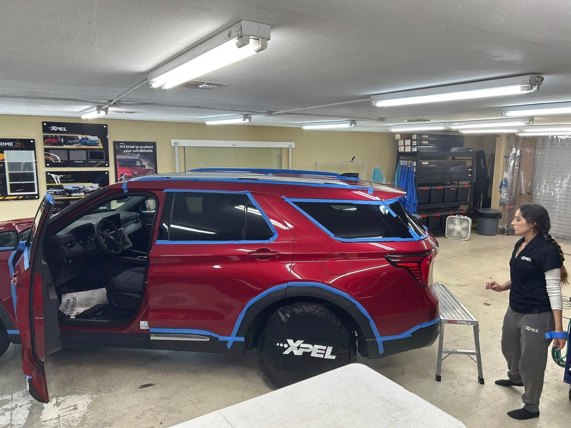 Red SUV in shop with blue tape outline, window tinting in progress. Woman watches, shop interior.