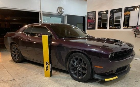 A plum Dodge Challenger parked inside, with a yellow post next to it. The tires and rims are black.