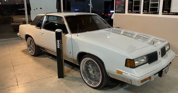 White classic Oldsmobile Cutlass with wire wheels parked near a building at night.