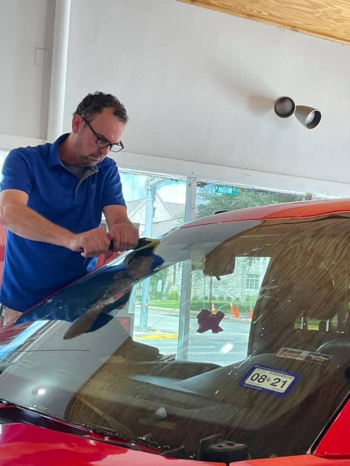 Man applies window tint to a red car windshield. He wears glasses and a blue shirt, working in a shop.