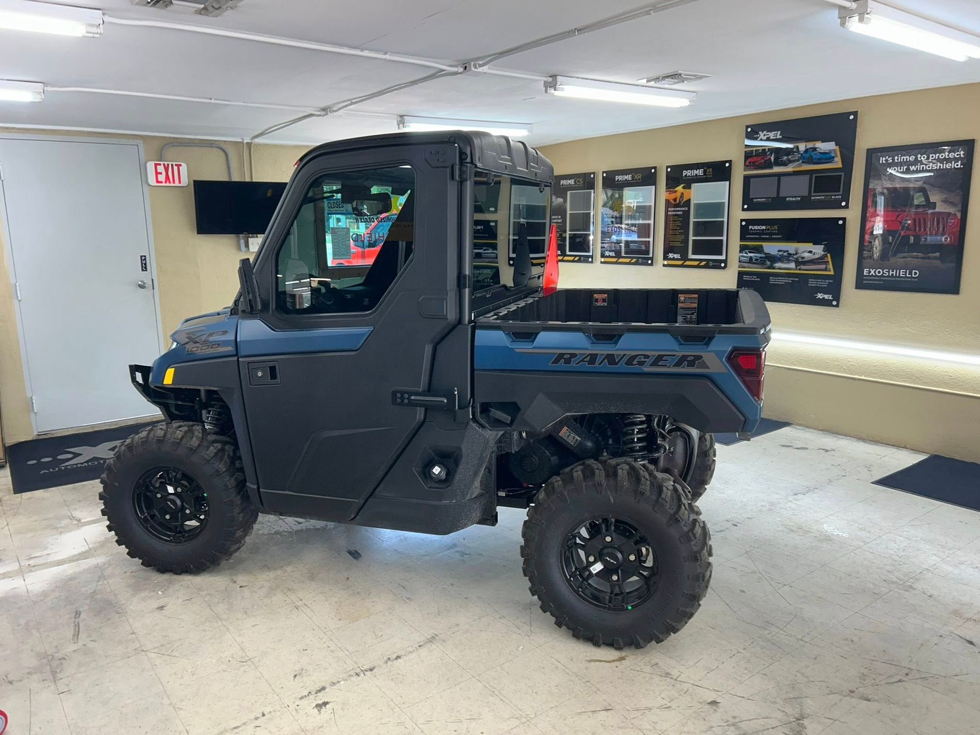 Blue and black Ranger side-by-side vehicle with large tires, inside a showroom with informational posters.