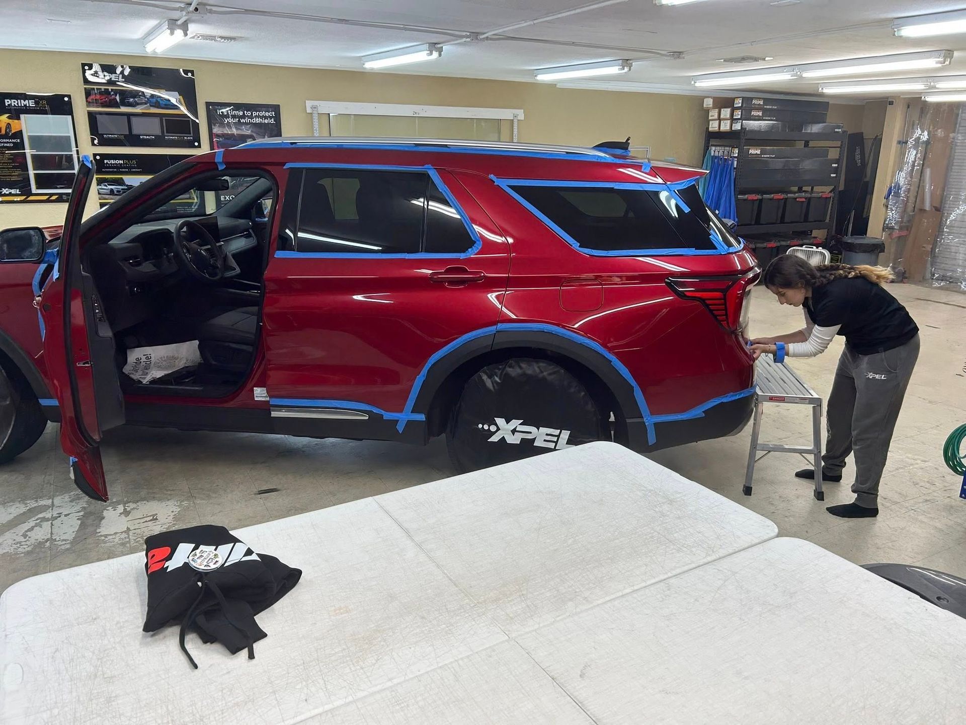Person working on a red SUV in a garage. Blue tape outlines panels; open door; white table in front.
