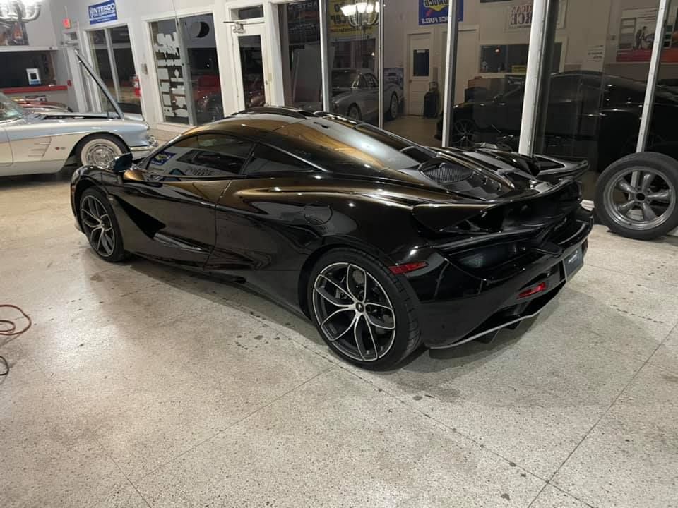 Black McLaren sports car parked indoors with a white car in the background.